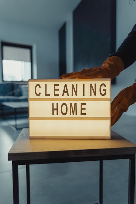 A person wearing orange cleaning gloves is adjusting a backlit sign on a table that reads 'CLEANING HOME', situated in a modern, well-lit room with a large window, light-colored tiled flooring, and a