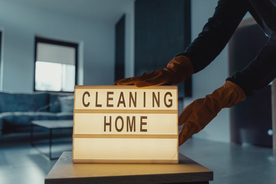 A person wearing orange cleaning gloves is adjusting a backlit sign on a table that reads 'CLEANING HOME', situated in a modern, well-lit room with a large window, light-colored tiled flooring, and a