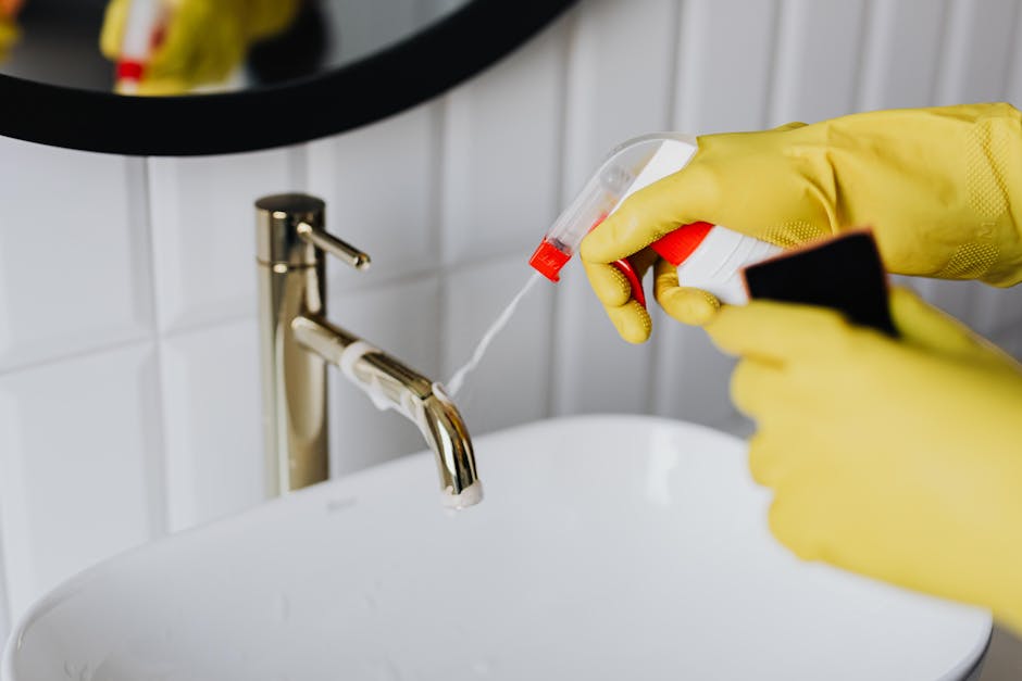 A professional cleaner from Bromley Cleaners wearing a white protective suit, blue gloves, and a face mask is using a handheld vacuum cleaner to deep clean a white sofa cushion in a bright living room